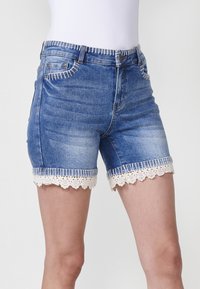 Džinsu šorti ar gaiši zilu mazgāšanu, pieguļošu dizainu un viļņotu mežģīņu apdari apakšmalā ar kontrastējošām baltām vīlēm.