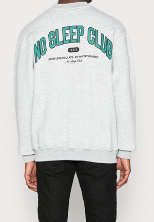 Hellgrauer Pullover mit grünem und schwarzem Schriftzug "NO SLEEP CLUB", getragen von einer Person, die mit dem Rücken zur Kamera steht, kombiniert mit schwarzer Hose.