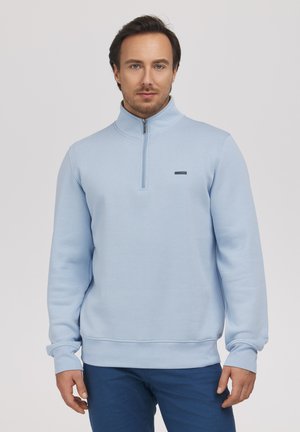 Mężczyzna w jasnoniebieskim pulowerze z półzamkiem i ciemnoniebieskich spodniach, stojący na tle jednolitego jasnego tła.