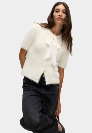 Hvid fluffy cardigan med korte ærmer, frontknaplukning og en stor hvid sløjfe ved halsudskæringen, parret med sorte jeans.
