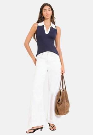 Frau trägt ärmelloses marineblaues Oberteil mit weißem Kragen, weiße Hosen mit weitem Bein, schwarze Sandalen mit Absatz und hält eine große braune Wildleder-Tasche.
