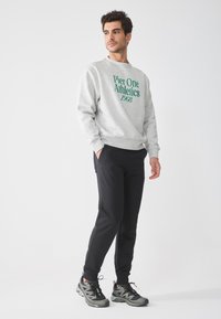 Tricou gri cu imprimație verde "Pier One Athletics 1968", asociat cu pantaloni sport negri și adidași gri-negru, stând în fața unui fundal alb.