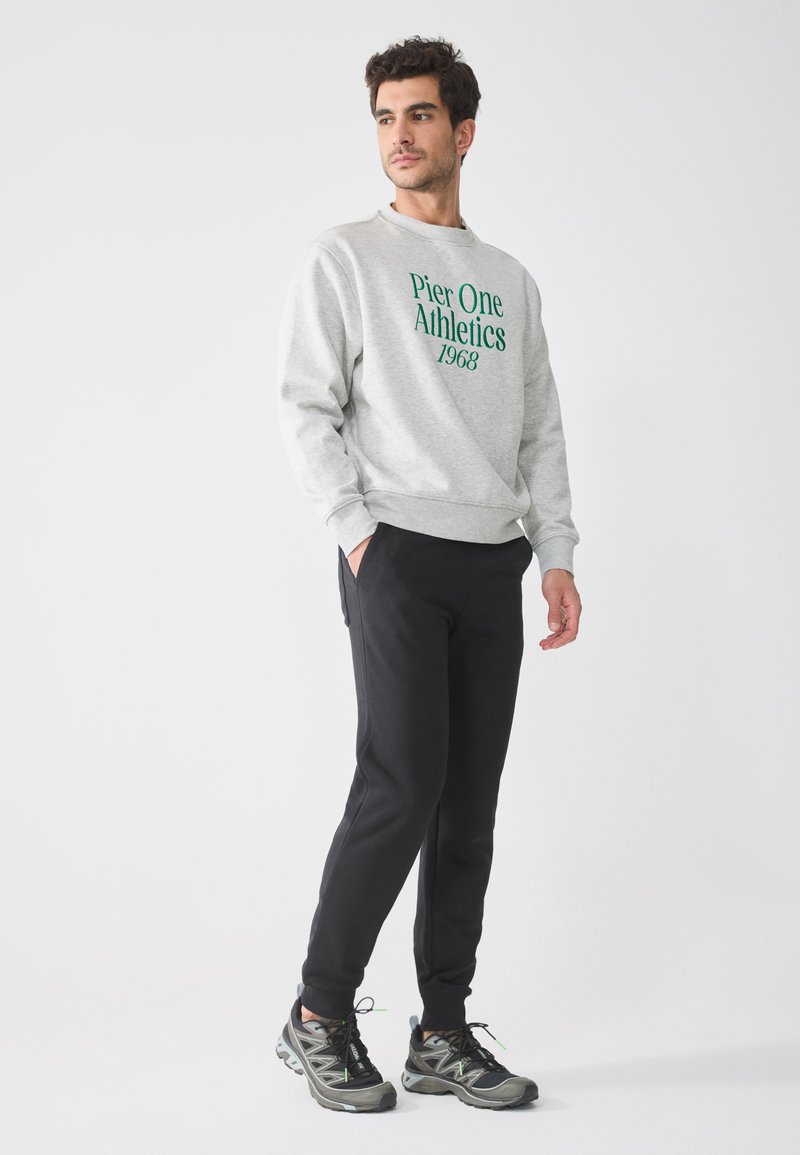 Tricou gri cu imprimație verde "Pier One Athletics 1968", asociat cu pantaloni sport negri și adidași gri-negru, stând în fața unui fundal alb.