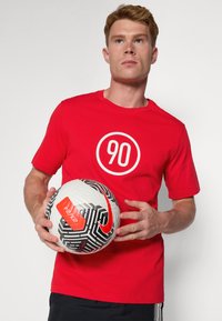 Rode katoenen T-shirt met een witte cirkelvormige graphic met het nummer 90. De persoon houdt een witte en zwarte gestructureerde voetbal met rode accenten vast.