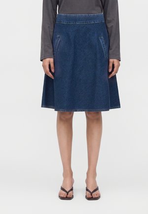 Femme portant une jupe en denim bleu foncé longueur genou avec des poches latérales, un haut gris à manches longues et des sandales noires à bout ouvert.