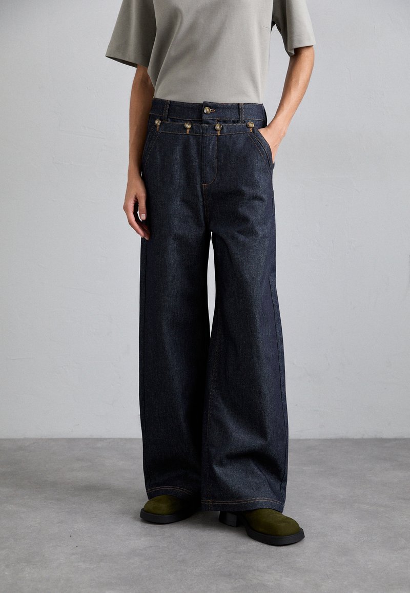 Pantalons en denim foncé à jambes larges avec des accents de boutons dorés, des coutures orange contrastantes, des poches latérales et une texture lisse, associés à des chaussures vert olive.