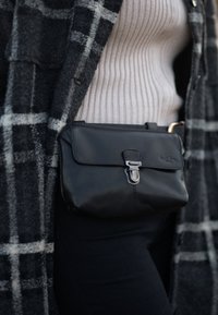 Černá kožená kabelka přes rameno s klopou a kovovým zapínáním. Vyznačuje se hladkým povrchem a jemným brandingem. Nosená přes žebrovaný krémový top.