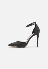 Schwarzer High Heel mit spitzem Zeh, Knöchelriemen mit goldenem Schnallenverschluss und strukturiertem Fersenbereich vor weißem Hintergrund.