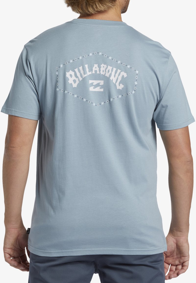 Tyrkysové bavlněné tričko má na zádech bílou grafiku se slovem "BILLABONG" a designem vlny, doplněnou dekorativními akcenty.
