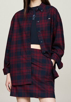 Femme portant une chemise oversize à carreaux rouges et bleu marine avec une jupe assortie et un top court noir, main dans la poche, sur fond beige.
