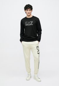 Schwarzes Langarmhemd mit weißem "EA7 Emporio Armani" Logo, kombiniert mit cremefarbenen Joggerhosen mit seitlichem Logo. Abgerundet wird das Outfit durch weiße Sneaker.