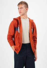 Narančasta zip-up dukserica s kontrastnim plavim trakama, koja ima prednji džep i rebraste manšete, nosi se preko svijetle bež majice i tamnih hlača.
