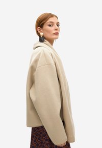 Mujer con abrigo beige de gran tamaño, grandes pendientes negros y dorados, y falda estampada marrón y azul marino, mirando de lado contra un fondo liso.