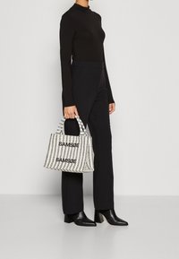 Eine schwarze, langärmlige Bluse und Hosen werden getragen, während eine gestreifte Tasche mit dem Wort "SAMSDØE" in Schwarz gehalten wird. Die Tasche hat zwei kurze Griffe.