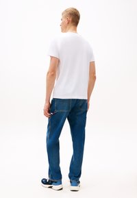 Biała krótka koszulka z krótkim rękawem, niebieskie dżinsy z detalami szwów oraz czarne buty sportowe z niebieskimi akcentami. Jednolite tło, widok na strój od tyłu.