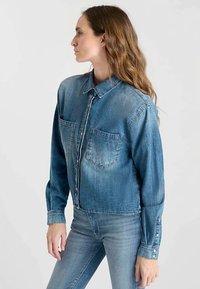 Cropped denim shirt in medium blue featuring a pointed collar, two chest pockets, button front, and long sleeves with button accents.