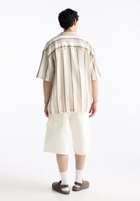 Camicetta a maniche corte a righe in beige e marrone, abbinata a pantaloni cargo bianchi. Tessuto texture, vestibilità casual, vista posteriore mostra tasche visibili.