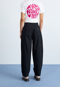 Witte t-shirt met roze cirkelvormig logo op de achterkant, gecombineerd met zwarte wijde broek en chunky, donkerbruine schoenen. Achtergrond met vlakke oppervlakte.