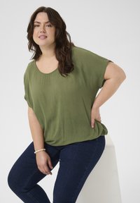 Mujer con cabello castaño ondulado, vestida con una blusa suelta de color verde oliva, jeans azul oscuro y una pulsera de perlas, sentada sobre un bloque blanco contra un fondo claro.