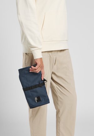 Pochette blu navy realizzata in tessuto testurizzato, con una tracolla nera e un logo, tenuta in mano, con una felpa color crema e pantaloni beige visibili.