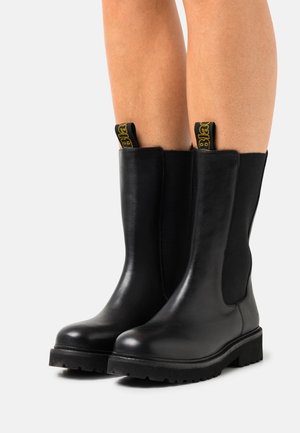 Schwarze Lederstiefel bis zur Mitte der Wade mit dicken Gummisohlen, getragen an nackten Beinen vor einem schlichten hellen Hintergrund.