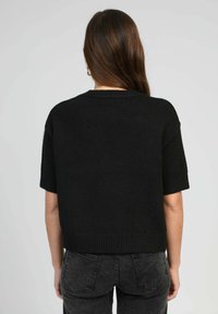 Pull en maille noir à manches courtes avec une coupe ample, ourlet côtelé et texture douce. Porté avec un jean gris, vu de l'arrière.