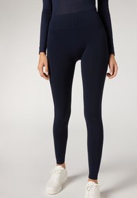 Mørkeblå højtaljede leggings lavet af strækbart stof med en glat overflade, tætsiddende form og ankellængde, parret med hvide sneakers.