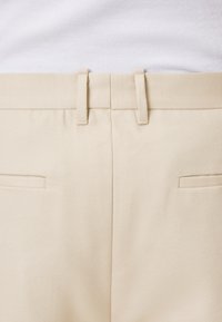 Pantalones de algodón beige con una textura suave, que cuentan con un frente plano, dos bolsillos traseros y pasadores para cinturón. Diseño simple y funcional.