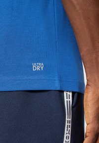 Plava košulja kratkih rukava s oznakom "ULTRA DRY" na rubu, izrađena od glatkog materijala. U kombinaciji je s tamnoplavim hlačama koje imaju bijelu prugu.