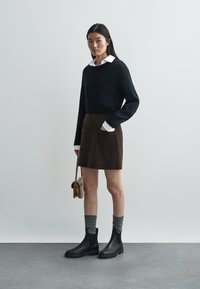 Melns džemperis, brūna velūra svārki ar divām priekšējām kabatām, pelēkas potīšu zeķes un melnas Čelsijas zābaki. Vienkārša soma papildina izskatu.