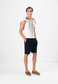 Junger Mann steht mit Händen in den Taschen, trägt ein weißes ärmelloses Shirt, marineblaue Shorts, einen grauen Pullover, der über den Schultern liegt, und braune Slipper.