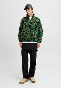 Pull en polaire vert et noir à motifs avec un col montant, associé à un pantalon noir et des chaussures beiges. Le modèle porte une casquette grise.