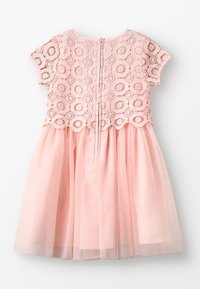 Jasnoróżowa sukienka z koronkowym topem ozdobionym okrągłymi wzorami i falowanym brzegiem, przechodząca w zwiewną spódnicę z tiulu. Zapinana na zamek z tyłu.