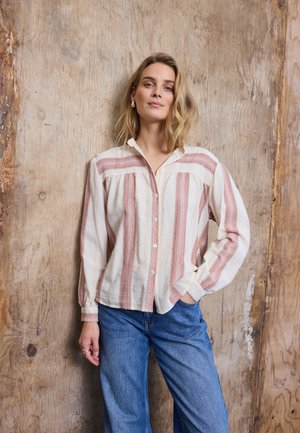 Hullámos szőke hajú nő áll egy fa fal előtt, csíkos krém és piros blúzt, valamint kék farmert visel, egyik keze zsebre téve.