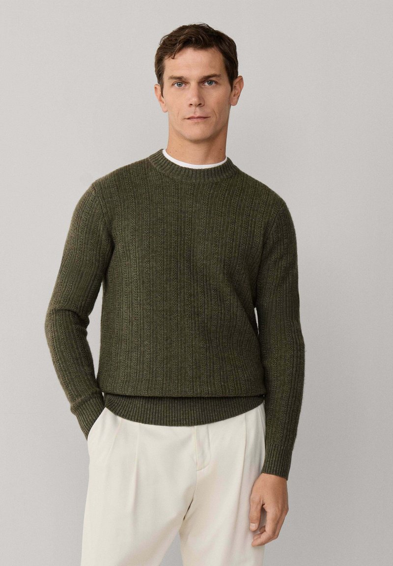 Maglione in maglia coste verde oliva con scollo rotondo, vita aderente e maniche lunghe; indossato sopra una camicia bianca e abbinato a pantaloni di colore chiaro.
