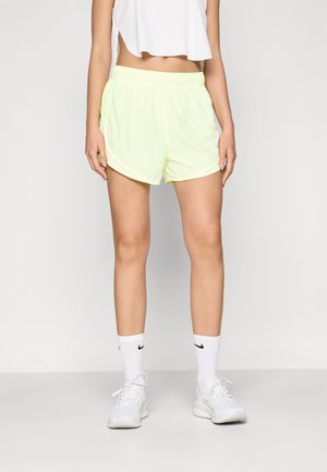 Personne portant un short de sport jaune clair, un top court blanc, des chaussettes Nike blanches et des baskets blanches, debout devant un fond uni.