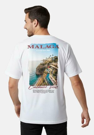 Man draagt een wit t-shirt met een afbeelding van een kustklif met palmbomen, oceaan en gebouwen, gelabeld met "Malaga Costa del Sol".