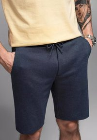 Pantaloni shorts azzurri con texture e vita regolabile tramite cordino, con tasche laterali e orlo sopra il ginocchio. Il materiale sembra resistente e leggero.
