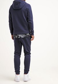 Tmavě modrá mikina s kapucí a odpovídající tepláky s texturovaným povrchem a elastickými manžetami. Spárováno s vzorovanou vnitřní vrstvou.