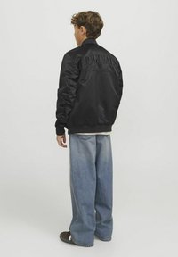 Svart bomberjacka med broderad "ORIGINALS" text på ryggen, ribbade ärmuddar och midja; bärs över en ljus skjorta och lösa denimjeans.