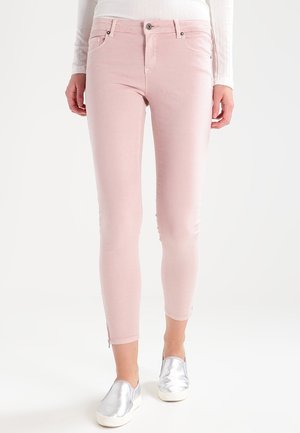 Pantalon slim rose clair porté avec des chaussures argentées slip-on et un haut blanc à manches longues côtelé sur une personne debout.