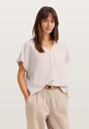 Woman with straight brown hair wearing a loose white V-neck top and beige pants with a tan belt, hands in pockets, neutral background.
