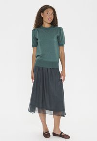 Zelena pletena majica s kratkimi napihnjenimi rokavi, v kombinaciji s dolgo, temno sivo polprosojno krilo. Model nosi preproste rjave balerinke.