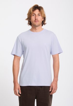 Homme aux cheveux mi-longs portant un t-shirt violet clair à manches courtes et un pantalon foncé, debout devant un fond blanc uni.
