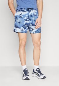 Bañador azul con patrones que presentan escenas tropicales, cintura con cordón y material elástico. Llevado con calcetines blancos y zapatillas navy.