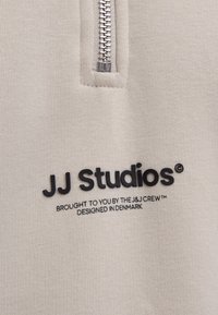 Beige vetoketjupaita, jossa on metallivetoketju, teksturoitu kangas ja musta painettu logo "JJ Studios" lisäbrändäys tekstillä alla.