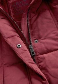 Bordeaux isoleret jakke med quiltet tekstur, udstyret med en metallynlås og trykknapper, mærket med branding og plejeinstruktioner.