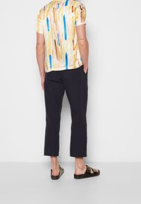 Man med kortärmad skjorta i flerfärgade vertikala ränder, svarta cropped byxor och svarta sandaler, står med händerna i fickorna och vänder sig mot väggen.