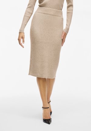 Kvinde iført en beige ribstrikket midi-nederdel med lange ærmer og sorte spidse ankelstropper, stående mod en hvid baggrund.