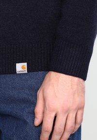 Granatowy sweterek dzianinowy z prążkowanymi mankietami, z małą białą i pomarańczową metką Carhartt. Materiał wydaje się miękki i teksturowany.